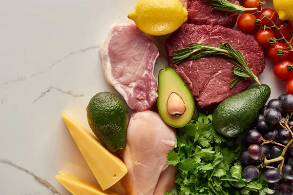 Vista superior de una variedad de carne cruda y aves de corral con aguacates, limón, queso, uvas, rama de tomates y ramitas verdes - foto de stock