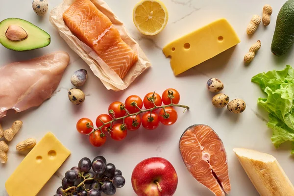 Top view of raw poultry and fish with avocados, lemon, cheese, eggs, grapes, cherry tomatoes, peanuts and apples — Stock Photo