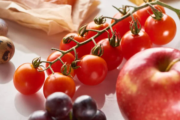 Vista de perto do ramo de tomates cereja perto de maçã, uva e ovos de codorna na superfície branca — Fotografia de Stock