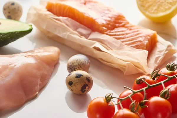 Vista de cerca del salmón crudo en papel pergamino y filete de aves cerca de huevos de codorniz, tomates, limón y aguacate en la superficie blanca - foto de stock