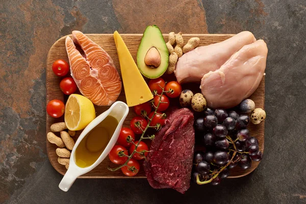 Top view of raw meat, fish, poultry, cheese, fruits, vegetables with olive oil and peanuts on wooden cutting board on dark brown marble surface — Stock Photo