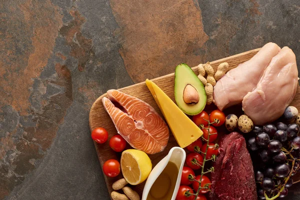 Vue de dessus de la planche à découper en bois avec du poisson cru, de la viande, de la volaille, du fromage, des fruits, des légumes, de l'huile d'olive et des arachides sur une surface en marbre brun foncé avec espace de copie — Photo de stock