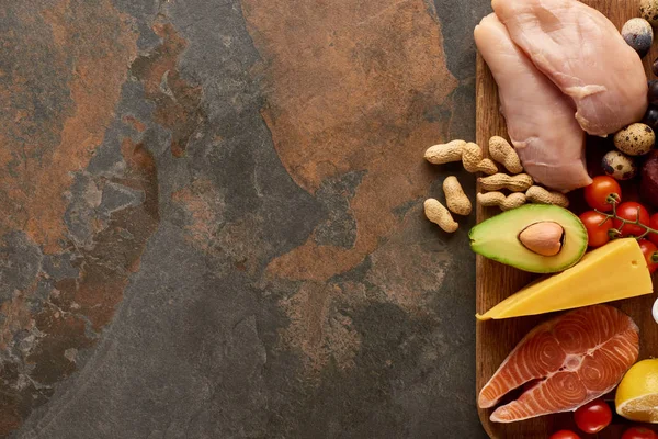 Top view of wooden cutting board with raw fish, poultry, cheese, fruits, vegetables, quail eggs, lemon and peanuts on dark brown marble surface with copy space — Stock Photo