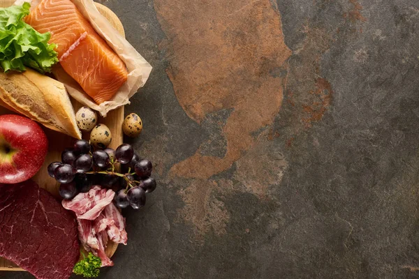 Top view of wooden cutting board with raw fish, meat, bacon, cheese, fruits, quail eggs, baguette and lettuce on dark brown marble surface with copy space — Stock Photo