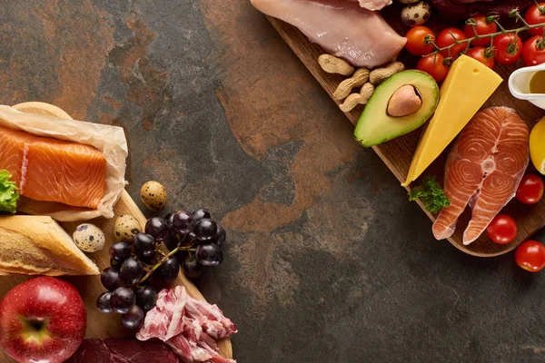 Vista superior de tablas de cortar de madera con pescado crudo, aves de corral, queso, frutas, verduras, tocino, aceite de oliva, baguette, huevos de codorniz y cacahuetes en la superficie de mármol marrón oscuro con espacio para copiar - foto de stock