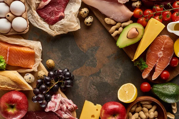Top view of wooden cutting board with raw fish, meat, poultry, cheese, fruits, vegetables, olive oil, eggs, baguette and peanuts on dark brown marble surface with copy space — Stock Photo
