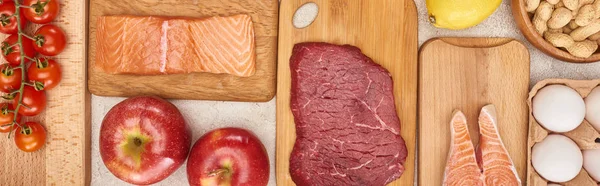 Panoramic shot of raw meat and salmon on wooden cutting boards near apples, eggs, tomatoes and peanuts — Stock Photo