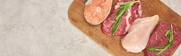 Panoramic shot of raw meat, fish and poultry with rosemary twigs on wooden cutting board on marble surface with copy space — Stock Photo