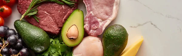 Panoramic shot of avocados, raw meat, chicken fillet, tomatoes, grape and cheese with greenery on marble surface — Stock Photo