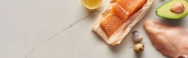 Panoramic shot of raw chicken breast near salmon on parchment paper, quail eggs, avocado and lemon on marble surface — Stock Photo