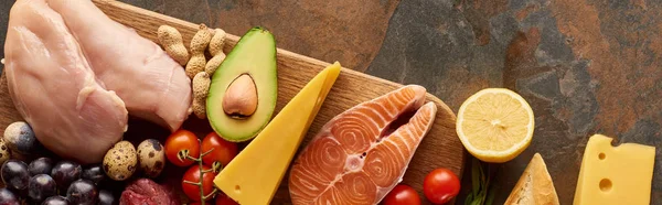Panoramic shot of wooden cutting board with raw fish and poultry near vegetables, grape, lemon, cheese and peanuts on marble surface — Stock Photo