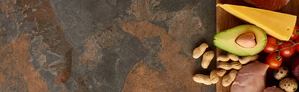 Tiro panorámico de tabla de cortar de madera con filete de aves de corral crudo, aguacate, tomates, queso, huevos de codorniz y cacahuetes en la superficie de mármol - foto de stock