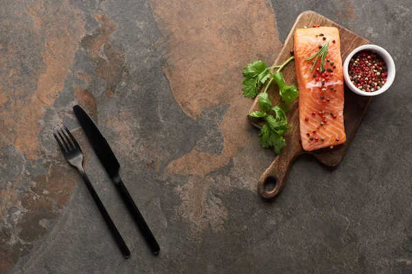 Vista superior de salmão fresco com pimenta, salsa em tábua de corte de madeira perto de talheres — Fotografia de Stock