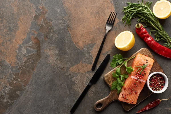 Top view of raw fresh salmon with herbs and lemon on wooden cutting board near cutlery and chili peppers — Stock Photo