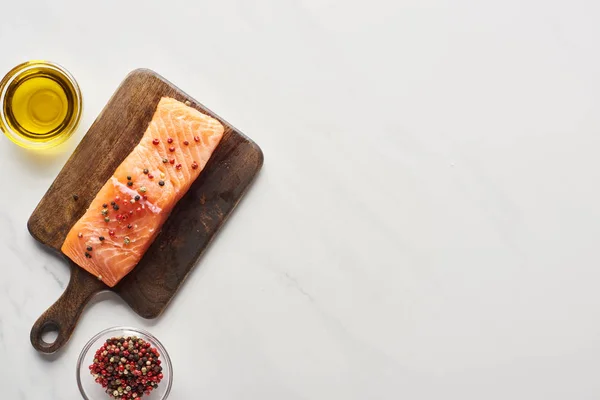 Top view of raw salmon steak with peppercorns on wooden cutting board near oil on marble surface — Stock Photo