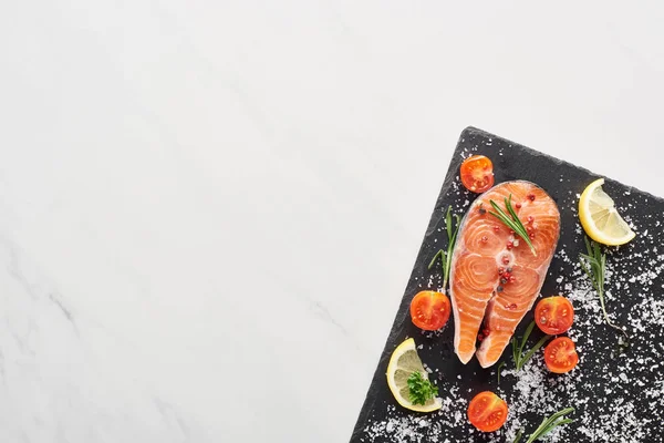 Вид сырого лосося с приправами и помидорами на каменной доске на мраморном столе — стоковое фото