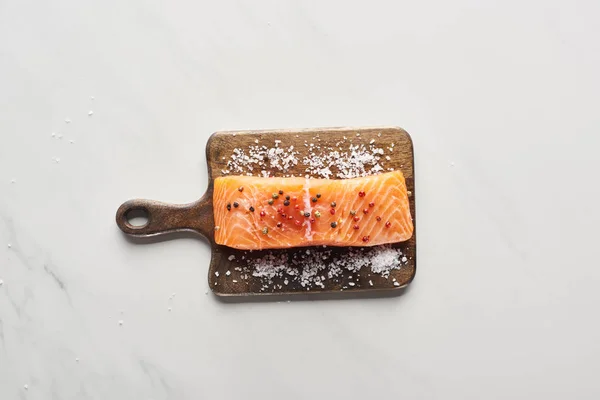 Top view of raw salmon steak with peppercorns and salt on wooden cutting board on marble surface — Stock Photo