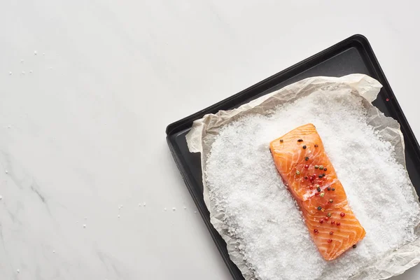 Вид сверху на стейк из сырого лосося с перцем на подносе с солью на мраморной поверхности — стоковое фото