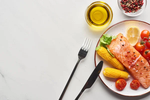 Vista superior del filete de salmón crudo con maíz y tomates en el plato cerca de cubiertos, aceite y granos de pimienta en la mesa de mármol - foto de stock