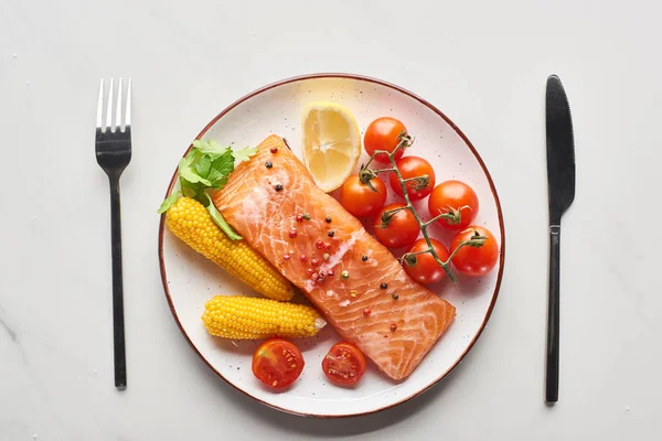 Top view of raw salmon steak served with corn and tomatoes on plate near cutlery on marble table — Stock Photo