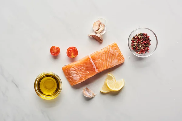 Top view of raw salmon steak near lemon, tomatoes, garlic, oil and peppercorns in bowls on marble table — Stock Photo