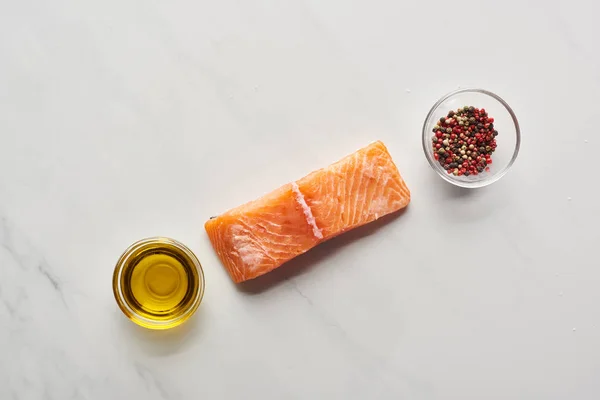 Vista dall'alto di bistecca di salmone crudo vicino all'olio e grani di pepe in ciotole sul tavolo di marmo — Foto stock