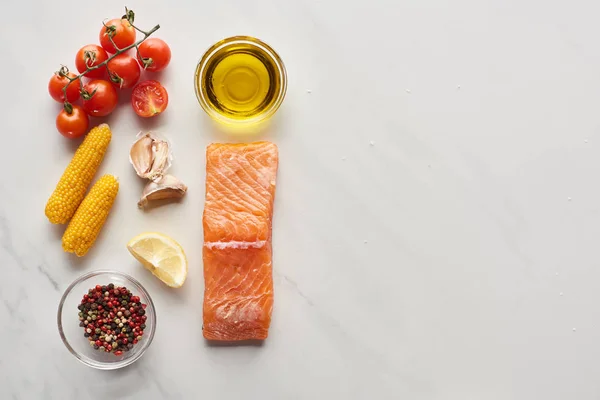 Vista dall'alto di bistecca di salmone crudo vicino a limone, mais, pomodori, aglio, olio e grani di pepe in ciotole sul tavolo di marmo — Foto stock