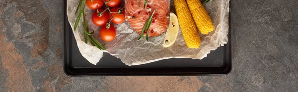 Panoramic shot of uncooked salmon steak with tomatoes, corn, rosemary, lemon and pepper on bakery paper on oven tray — Stock Photo