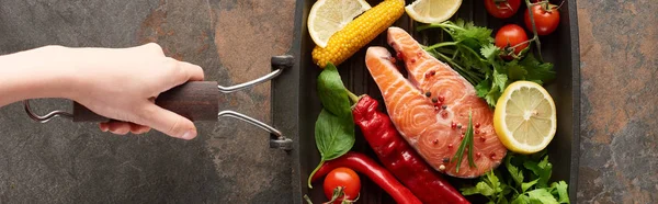 Partial view of woman holding raw salmon with vegetables, lemon and herbs in grill pan, panoramic shot — Stock Photo