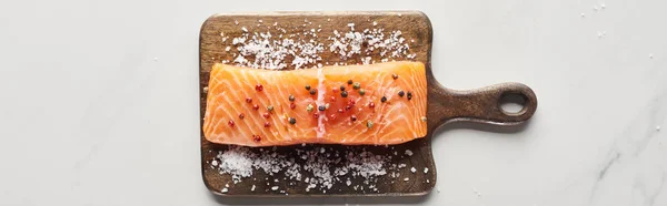 Top view of raw salmon steak with peppercorns and salt on wooden cutting board on marble surface, panoramic shot — Stock Photo