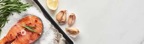 Top view of uncooked salmon steak with rosemary on oven tray with salt near garlic and lemon, panoramic shot — Stock Photo