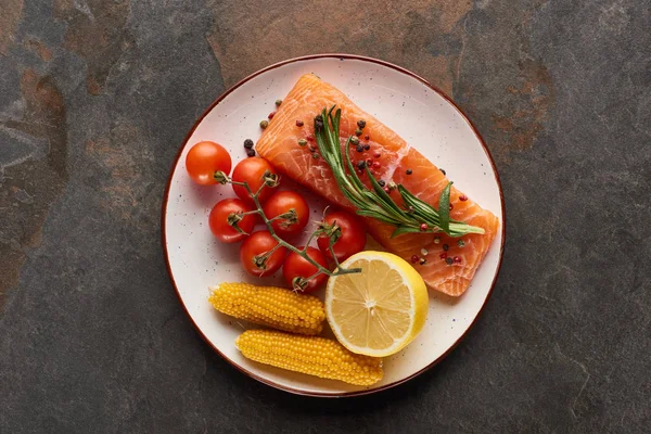 Вид сверху на сырой стейк лосося с помидорами, кукурузой, лимоном, розмарином и перцем на тарелке — стоковое фото