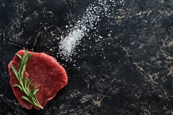 Top view of raw beef steak with rosemary twig on black marble surface with scattered salt crystals — Stock Photo