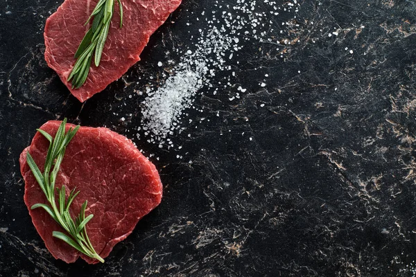 Vue de dessus des steaks de boeuf crus avec des brindilles de romarin sur la surface de marbre noir avec des cristaux de sel dispersés — Photo de stock