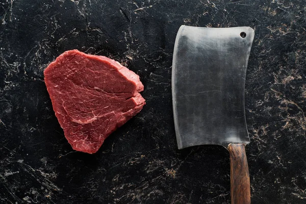 Top view of butcher knife near raw beef steak on black marble surface — Stock Photo