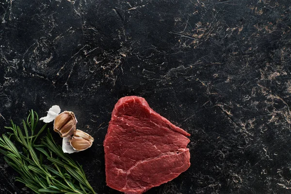 Vista superior de bife de carne crua perto de galhos de alecrim e dentes de alho na superfície de mármore preto — Fotografia de Stock