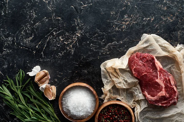 Vista dall'alto di parte di carne cruda su carta pergamena vicino a spezie, aglio e rosmarino su superficie di marmo nero — Foto stock