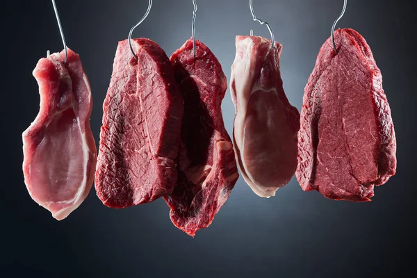 Assorted raw meat steaks on metal hooks on dark black background — Stock Photo