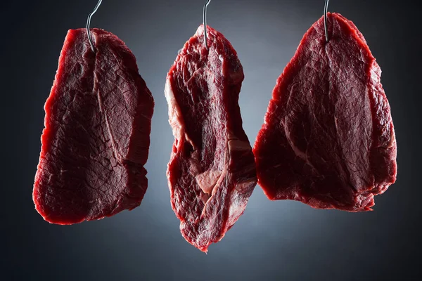 Raw beef steaks on metal hooks on dark black background — Stock Photo