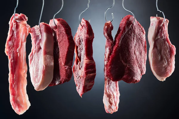 Assorted raw meat and bacon on metal hooks on dark black background — Stock Photo