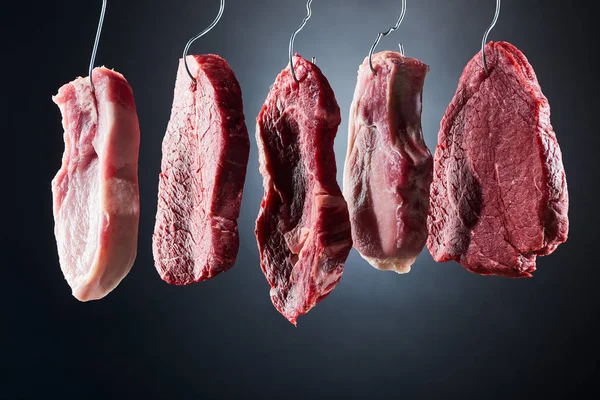 Assorted raw meat on metal hooks on dark black background — Stock Photo