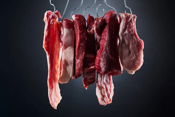 Bifes de carne crua sortidas e bacon em ganchos de metal no fundo preto escuro — Fotografia de Stock