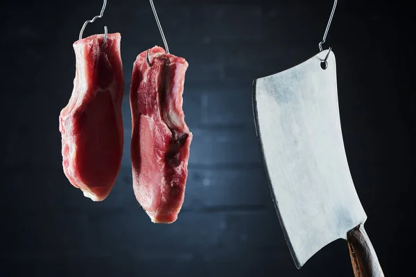 Filetes de cerdo crudos y cuchillo de carnicero en ganchos de metal sobre fondo negro oscuro - foto de stock