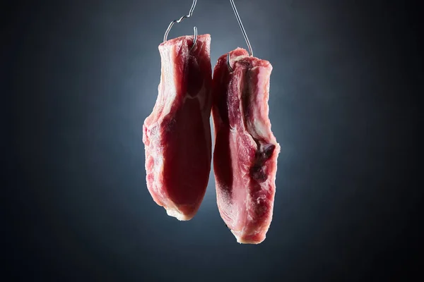 Raw pork pieces on metal hooks on dark black background — Stock Photo