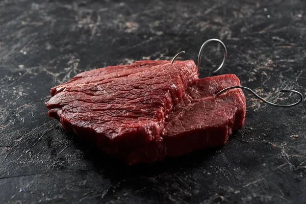 Bifes de carne crua com ganchos de metal na superfície de mármore preto — Fotografia de Stock