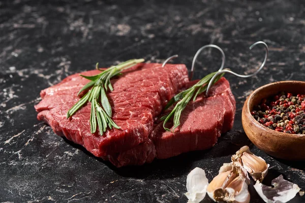 Bistecche di carne cruda con ramoscelli di rosmarino vicino a piccola ciotola con grani di pepe e aglio sulla superficie di marmo nero — Foto stock