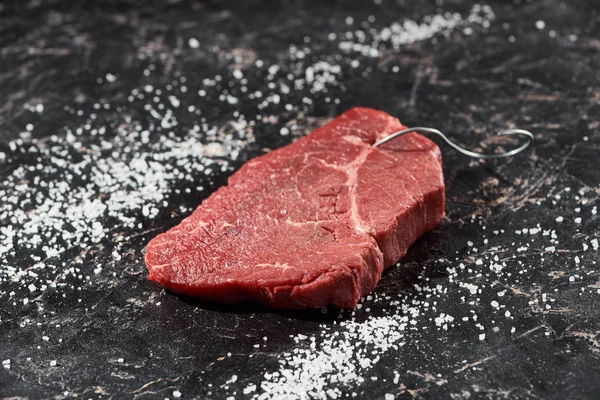 Raw meat steak with metal hook on black marble surface with scattered salt — Stock Photo