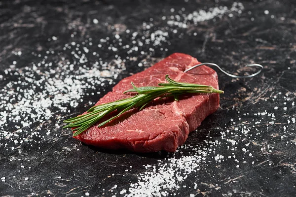 Raw meat steak with rosemary twig on black marble surface with scattered salt — Stock Photo