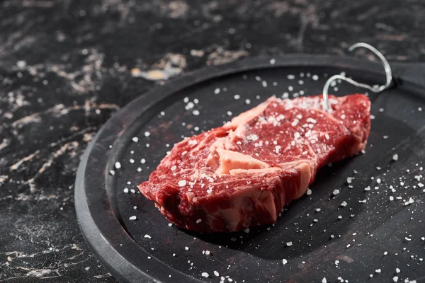 Carne crua polvilhada com sal e pimenta em placa de madeira na superfície de mármore preto — Fotografia de Stock