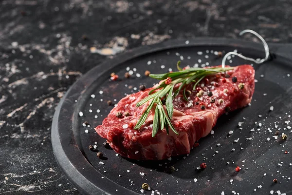 Steak de viande crue avec brindilles de romarin saupoudrées de sel et de poivre sur une surface ronde en bois — Photo de stock